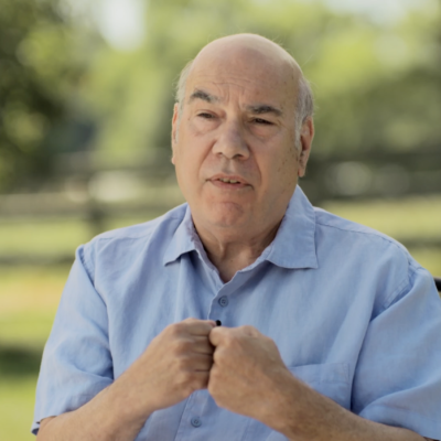 Jay Parini wears a light blue short sleeve shirt and gestures with his hands as speaks, sitting outside in a grassy garden.