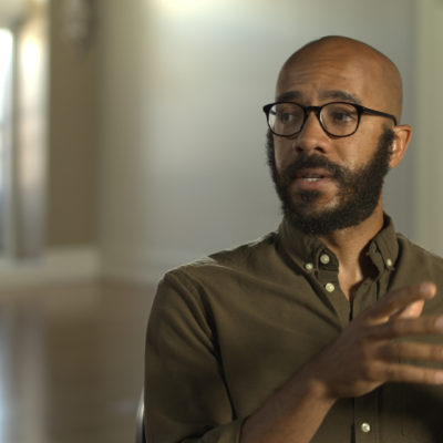 Clint Smith wears a dark green shirt and glasses, gesturing with his hands as he speaks.