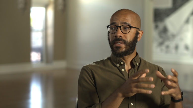 Clint Smith wears a dark green shirt and glasses, gesturing with his hands as he speaks.