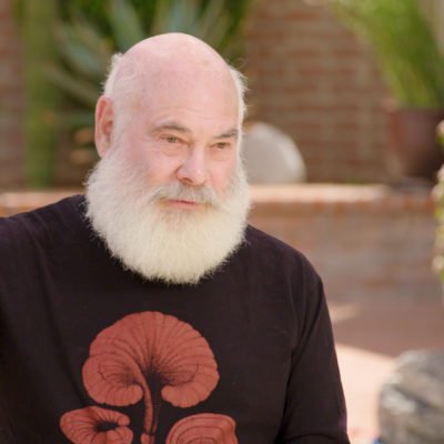 Dr Andrew Weil, wearing a black top with a red design, gestures with his hand as he speaks.