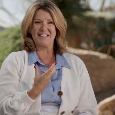 Paula Byrne, wearing a blue shirt and a white jackets, smiles and gestures with her hands as she speaks.