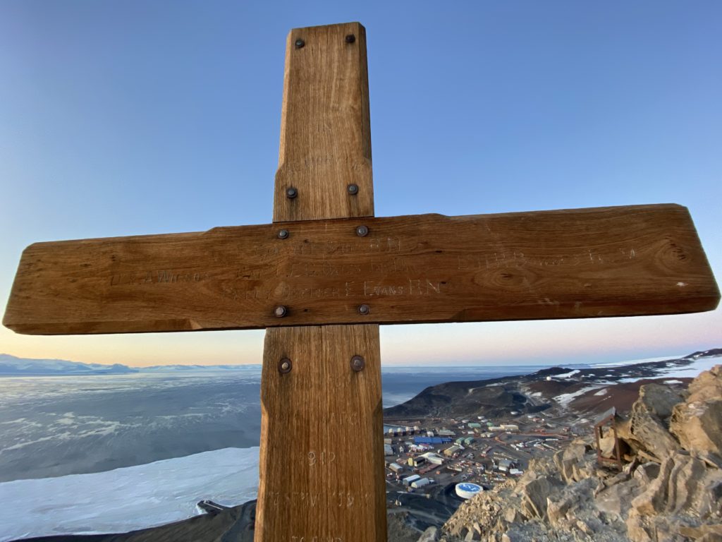 The wooden cross is against a bright blue sky, below it, at the bottom of the mountain, are some buildings.