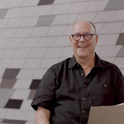 David Baker, wearing a black top, holds paper in his hand as he smiles.