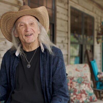 Jimmie Dale Gilmore, wearing a hat, gestures with his hand as he speaks.