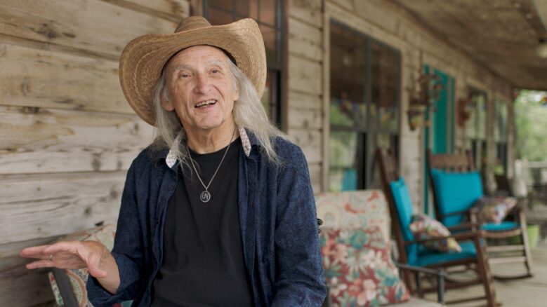 Jimmie Dale Gilmore, wearing a hat, gestures with his hand as he speaks.