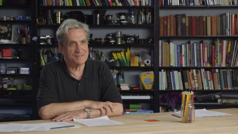 Robert Pinsky smiles while sitting with arms folded at a table