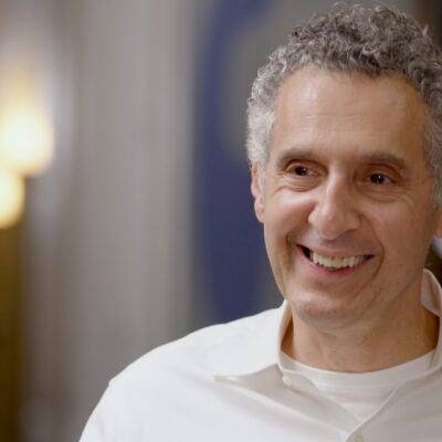 John Turturro smiles while wearing a white collared shirt