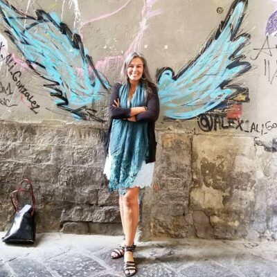 Lupita Eyde-Tucker smiles while crossing her arms in front of a wall with a winged graffiti