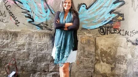 Lupita Eyde-Tucker smiles while crossing her arms in front of a wall with a winged graffiti